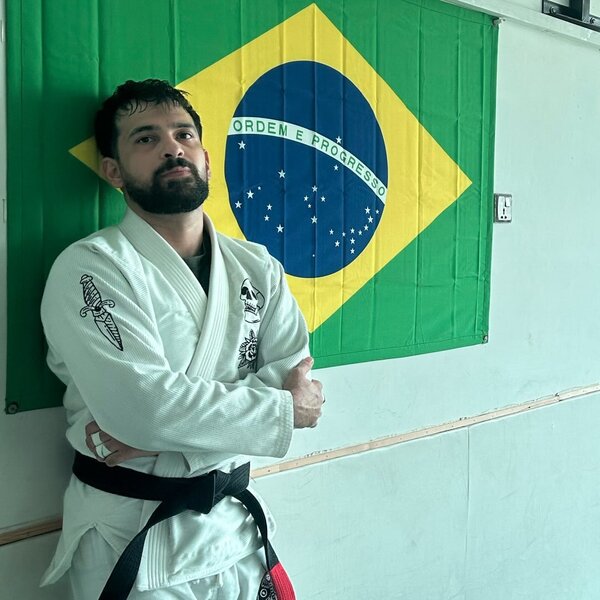 Shaheryar Khalid in a white gi and black belt, arms crossed in front of a Brazilian flag